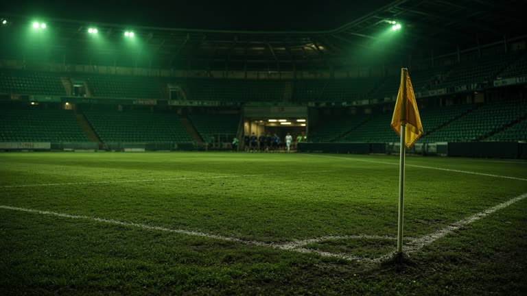 Empty football stadium corner under green floodlights with a corner flag and worn pitch grass, players blurred in the distant tunnel mouth, evening match atmosphere, no logos or readable text