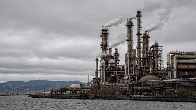 Industrial oil refinery towers and pipe racks beside San Francisco Bay under gray overcast sky, steam vents and weathered steel, distant hills, no logos or readable text