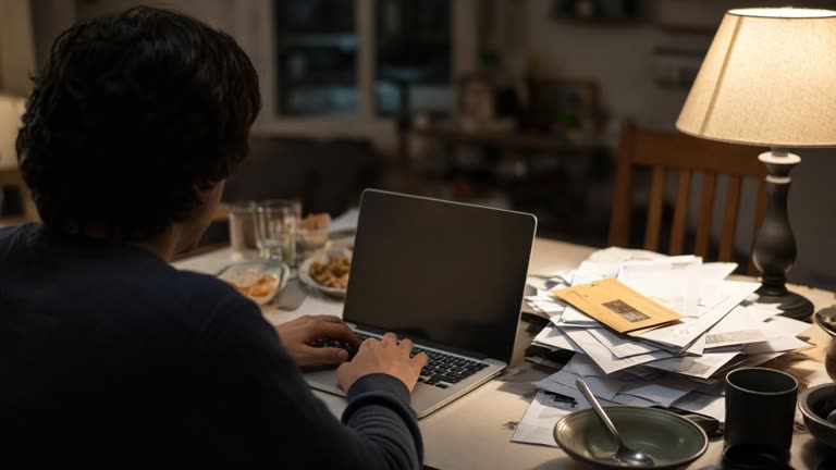 Consumer typing on a laptop at a cluttered dining table with mail piled nearby, evening lamp light, no logos or readable text