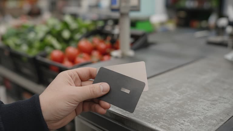 Close view of a hand holding two credit cards at a supermarket checkout lane, blurred produce in background, worn plastic counter, no logos or readable text