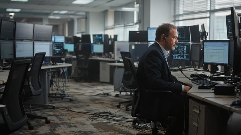 Securities firm trading floor with rows of monitors and a compliance officer wearing a wired headset at a side desk, overhead fluorescents and muted daylight through blinds, scuffed carpet and cable bundles, no logos or readable text