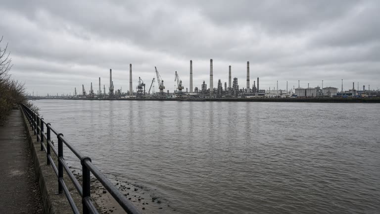 View from a Thames riverside walkway toward grey refinery stacks and cargo cranes on the horizon, overcast afternoon light, rippling water, no logos or readable text