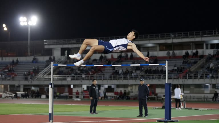 Taiwanese high jumper Fu Chao-hsuan in white and blue uniform clearing the bar at the Nagoya Asian Games stadium with officials and a sparse night crowd in the background
