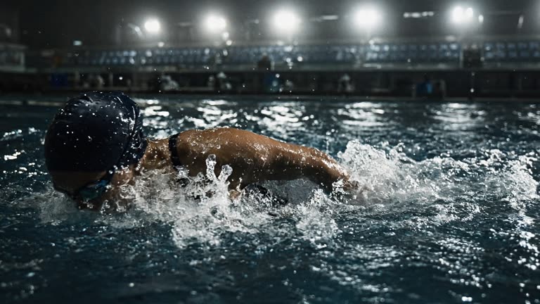 A female competitive swimmer in a dark cap and racing suit mid-stroke in an outdoor 50m pool under stadium floodlights, water churning white at her shoulder, blurred lane ropes and half-empty grandstand seating behind, shot from a low poolside angle with a long lens in cool evening light, no logos or readable text