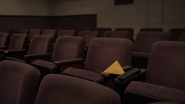 Small arthouse cinema screening room with rows of empty seats and a folded golden envelope on a side table beside a television remote on worn velvet upholstery, dim aisle lighting, no logos or readable text