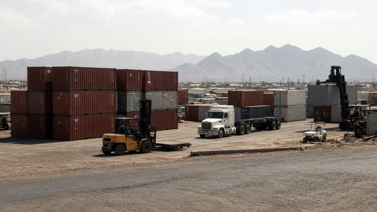 Industrial container yard beside a highway in northern Mexico at midday, stacked shipping crates and forklifts, hazy mountains in the distance, harsh sunlight, dusty asphalt, no logos or readable text