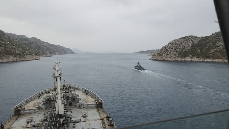Hazy midday view from a tanker bridge toward a narrow blue strait flanked by rocky coastlines, escort patrol boat trailing in the distance, heat shimmer on the water, no flags or readable hull markings