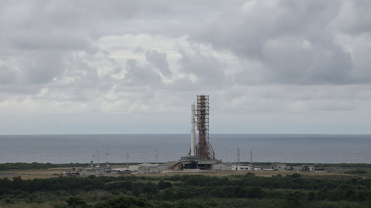 Rocket on a launch pad at Tanegashima Space Center viewed from a distance, ocean horizon and cloudy sky, no logos or readable text