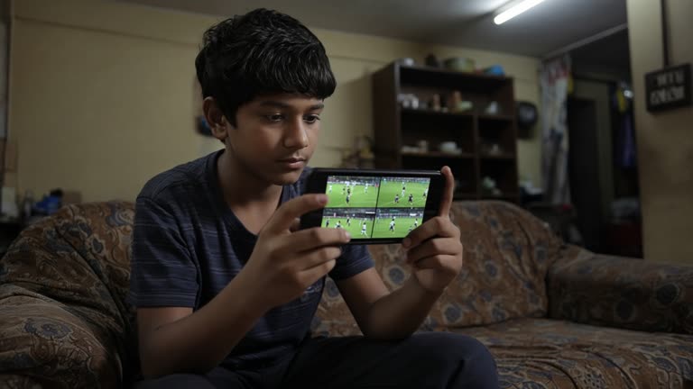 Young viewer holding a smartphone showing four small live sports video tiles on a couch in a modest Indian living room, evening tube light, worn upholstery, no readable screen text or logos