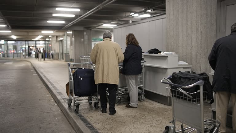 Domestic airport terminal curbside with a small assisted-travel desk and wheeled luggage carts under bright ceiling lights, faces turned away, no logos or readable text