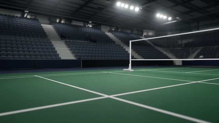 Indoor badminton arena with green court and empty spectator stands under bright overhead lights, low angle documentary sports photo, no faces, no logos or readable text