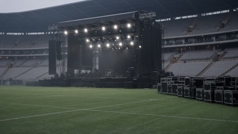 Outdoor stadium concert stage under rigging lights with empty seats and road cases stacked at the sideline, dusk haze over the field, no logos or readable text