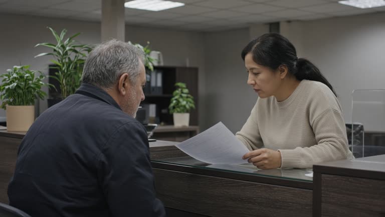 Bank customer reviewing insurance documents with a teller at a quiet service counter with potted plants and diffused ceiling light, forms turned away from camera, no logos or readable text