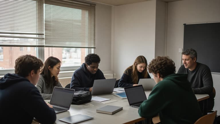 University seminar room with students and instructor around laptops, afternoon light through horizontal blinds, projector off, no readable text on screens, no logos