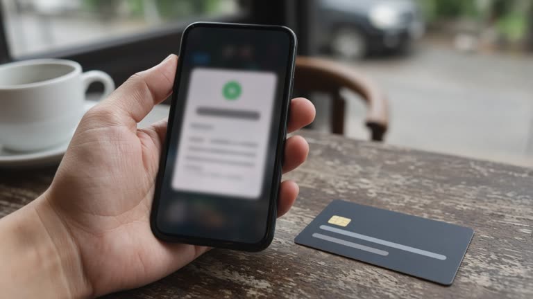 Hand holding a smartphone with a blurred banking notification on screen next to a debit card on a cafe table, daylight, no logos or readable text.