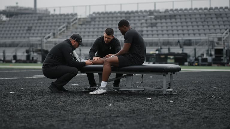 NFL team practice field with athletic trainers tending to a player seated on a bench, overcast morning light, rubber turf pellets visible, blurred stadium seats in background, no logos or readable text