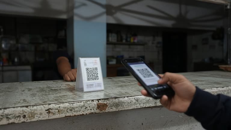 Small shop counter with a laminated UPI QR stand and a customer smartphone held at payment angle, worn Formica surface and ceiling fan shadows, no logos or readable text