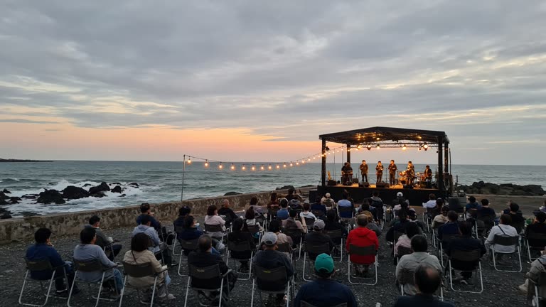 Photoreal wide shot of a lit open-air festival stage on a Penghu waterfront at dusk, rows of folding chairs filled with a seated crowd facing a low sea wall, string lights and lantern installations strung behind them, moderate surf breaking on dark rocks, overcast sky with the last warm band of light at the horizon; no logos or readable text.