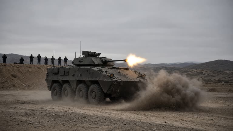Wheeled armored vehicle on dusty training range firing main gun toward distant hills, muzzle flash and dust plume, overcast sky, photographers on berm in background, documentary wire photo, slight underexposure