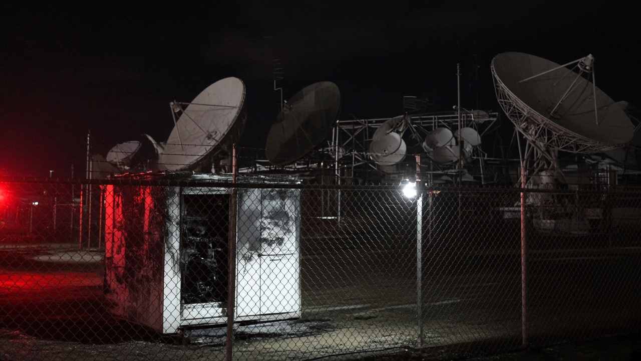 Satellite dish farm behind chain-link fence with scorched electrical cabinet and emergency lights, night scene, documentary wire photo, slight underexposure, no identifiable faces