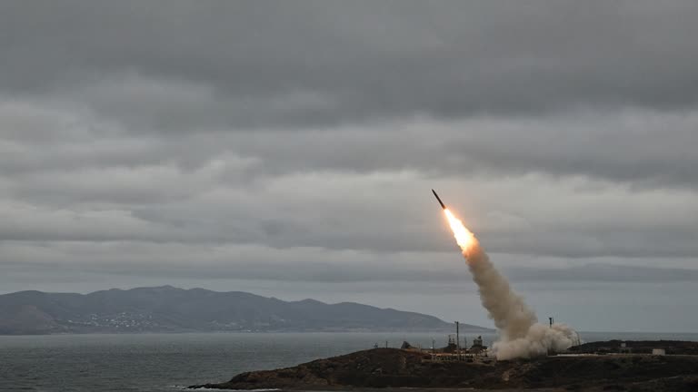 A short-range ballistic missile climbs from a coastal launch area on a gray afternoon, vapor trail against low clouds over the sea, distant hills, no logos or readable text.