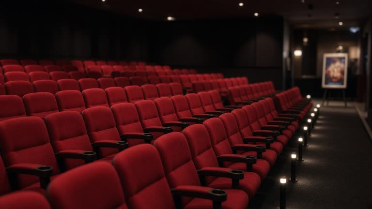 Empty multiplex auditorium with red fabric seats and dim aisle lighting, blurred standing poster easel at lobby entrance, carpet wear patterns, no logos or readable text