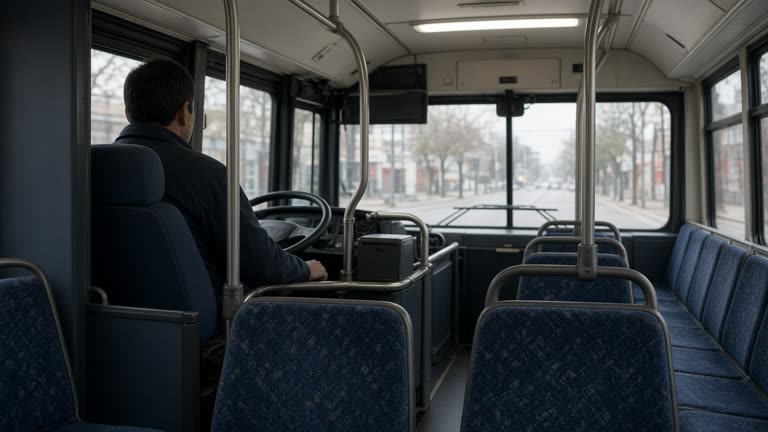 Bus captain at the wheel of a public bus seen from the aisle, fluorescent cabin light on empty fabric seats and stainless handrails, overcast daylight through windshield, no logos or readable text