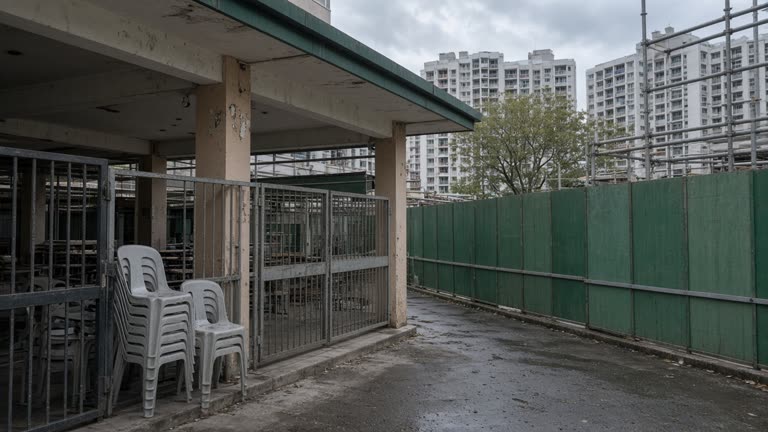 HDB hawker centre frontage with green renovation hoarding and scaffolding along the walkway, Clementi estate blocks in background, cloudy afternoon light, empty plastic chairs stacked inside gated entrance, no logos or readable text