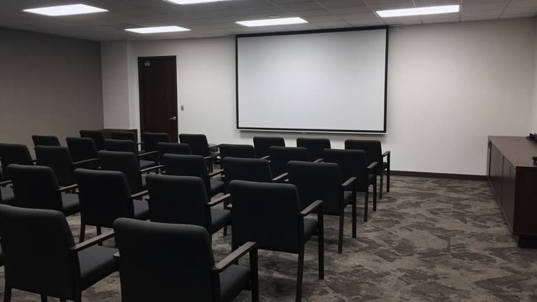 Empty government conference room with rows of chairs facing a blank projection screen, cool fluorescent ceiling panels and carpet wear patterns, no logos or readable text