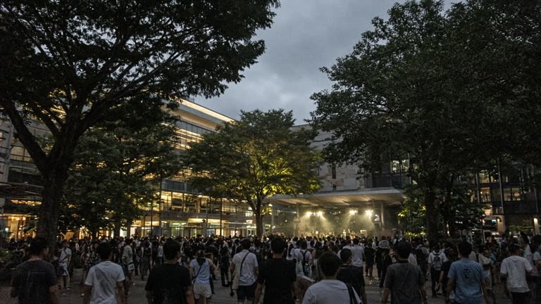 Orchard Road evening with crowd silhouettes and soft stage lighting on a civic plaza, trees and mall facades without readable signage, no logos or readable text
