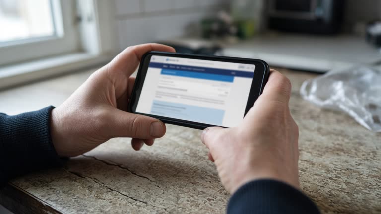 Close view of hands holding a smartphone over a kitchen table, blurred banking-style webpage on screen without readable text, daylight from window, worn laminate surface, no logos or readable text