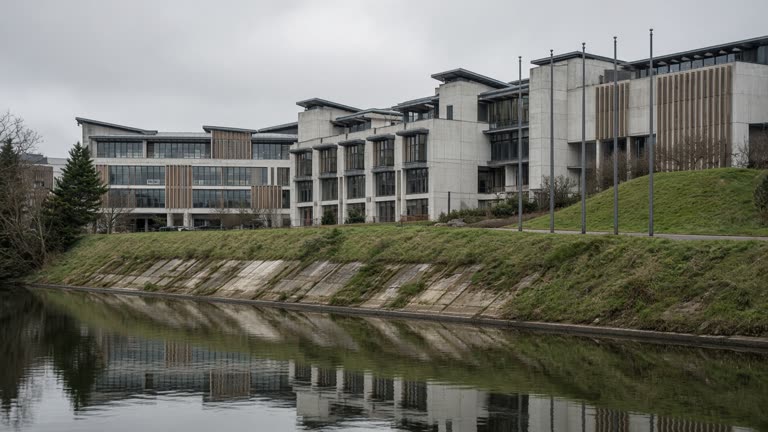 Scottish Parliament building exterior across calm water under grey sky, flagpoles without readable flags, concrete and grass banking, documentary framing, no logos or readable text