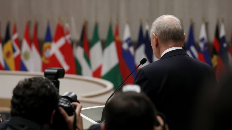 UN General Assembly rostrum with muted national flags out of focus behind a dark suit shoulder and microphone, photographers silhouetted in foreground, documentary handheld framing, no logos or readable text