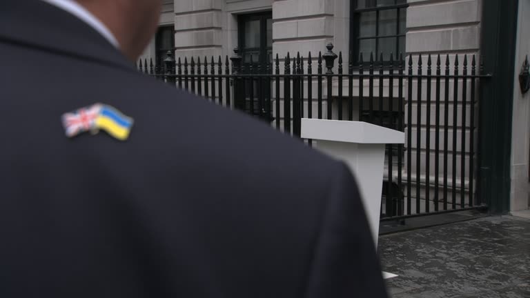 Downing Street podium with union flag and Ukraine flag pin on a dark suit jacket blurred in foreground, grey September daylight through black railings, no readable text or logos