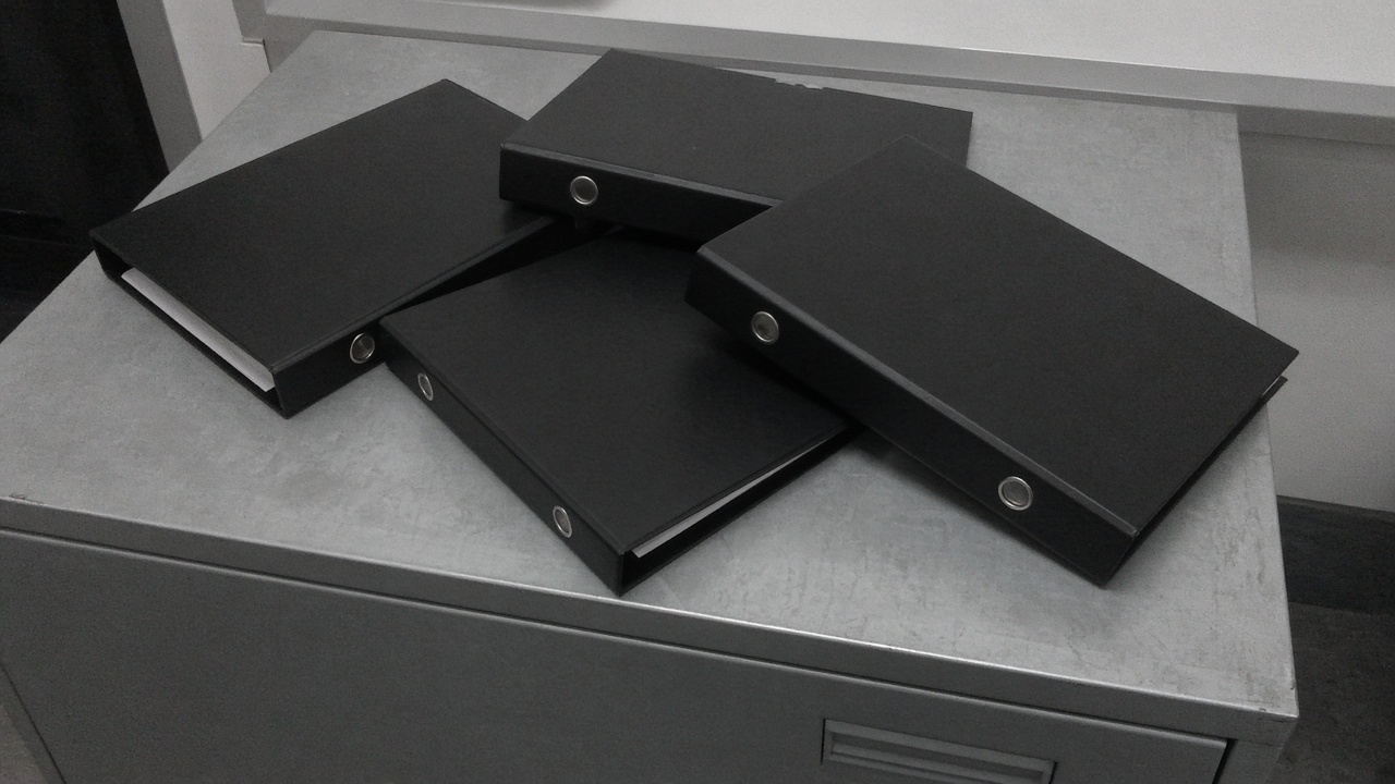 Two closed blank black ring binders on their sides on a wood table, overcast window light, no people, no logos, no readable text