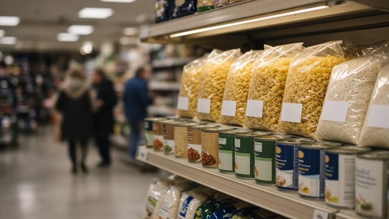 Supermarket shelf stocked with generic own-brand pasta, rice and tinned goods under warm strip lighting, blurred shoppers in background, no readable brand names or logos.