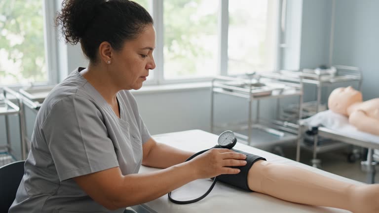 Adult learner at a college nursing skills lab practising blood pressure on a training mannequin, bright classroom windows and stainless steel trolleys, no logos or readable text