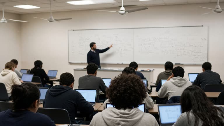 Undergraduate lecture hall with students on laptops during a morning seminar, projector glow on whiteboard diagrams, ceiling fans, no readable slides or logos