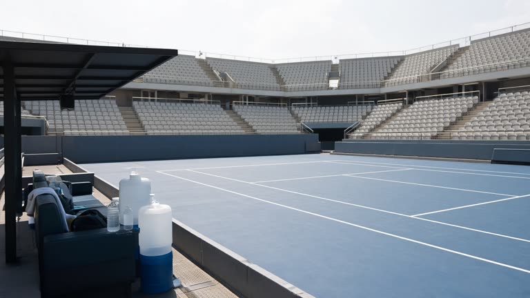 Hard tennis court at a large stadium with shaded player benches and water coolers at the sideline, bright midday sun and hazy sky, empty seats in background, no logos or readable text