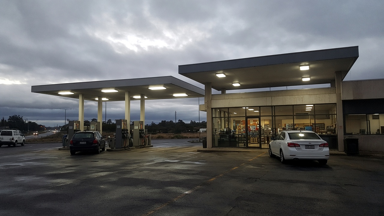 Australian highway fuel and convenience site at dusk with canopy lights on, cars at pumps and glass-front shop, overcast sky, worn asphalt, no logos or readable text
