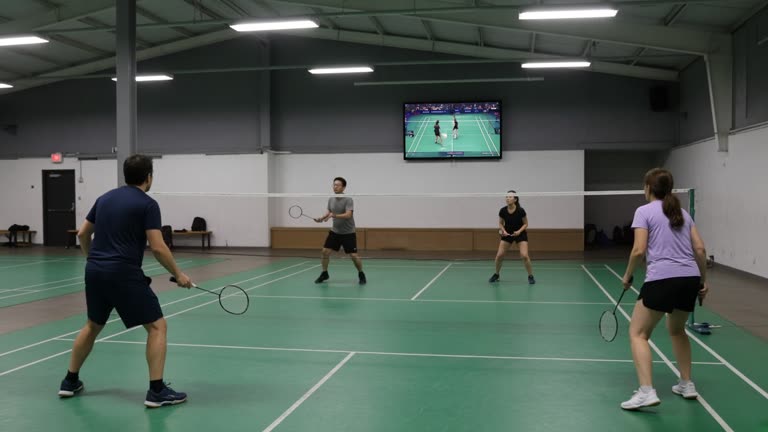Adults playing badminton on municipal community center courts with a wall-mounted television showing a sports broadcast in the background, fluorescent gym lighting and scuffed green flooring, no logos or readable text