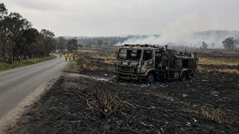 A blackened roadside on Katanna Road near Wedderburn in south-west Sydney after a hazard reduction burn escaped containment, with a destroyed fire truck on a dirt verge, emergency crews in the middle distance and smoke drifting over paddocks, photoreal, no logos or readable text.