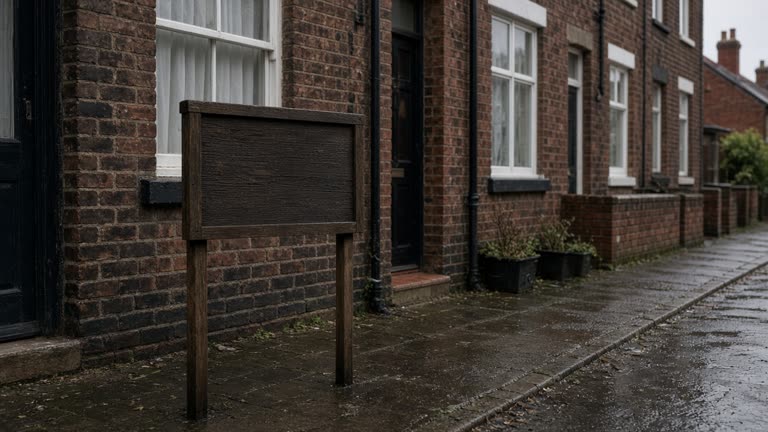 Row of brick terraced houses with a wooden for-sale sign on a damp pavement under overcast light, muted colours and slight grain, no logos or readable text