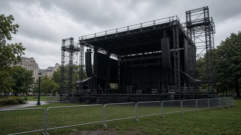 Outdoor concert stage scaffolding in a city park on a cloudy day, empty crowd barriers and wet grass, no logos or readable text