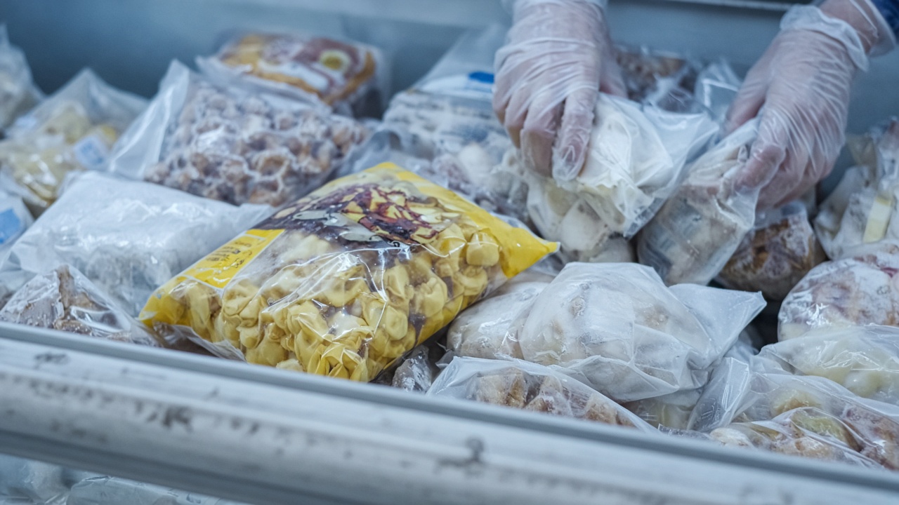 Yellow frozen pasta bag lying in a supermarket freezer case among frosted packages, cool blue lighting, shopper hands in gloves placing items, no readable labels or logos