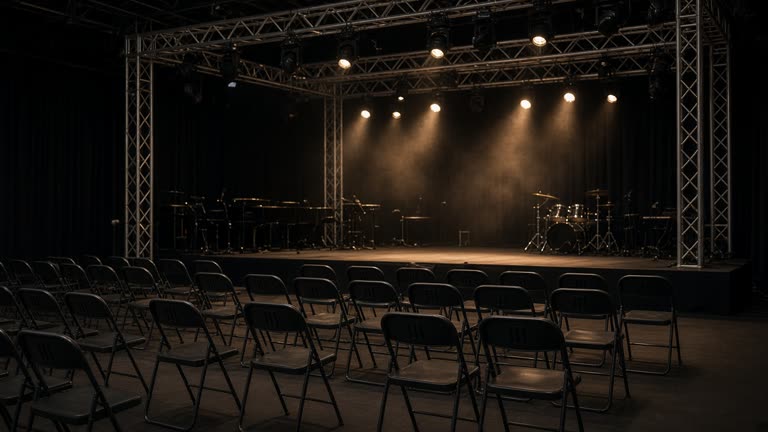 Indoor concert hall stage with truss lights and empty folding chairs before doors open, dim house lights, no logos or readable text