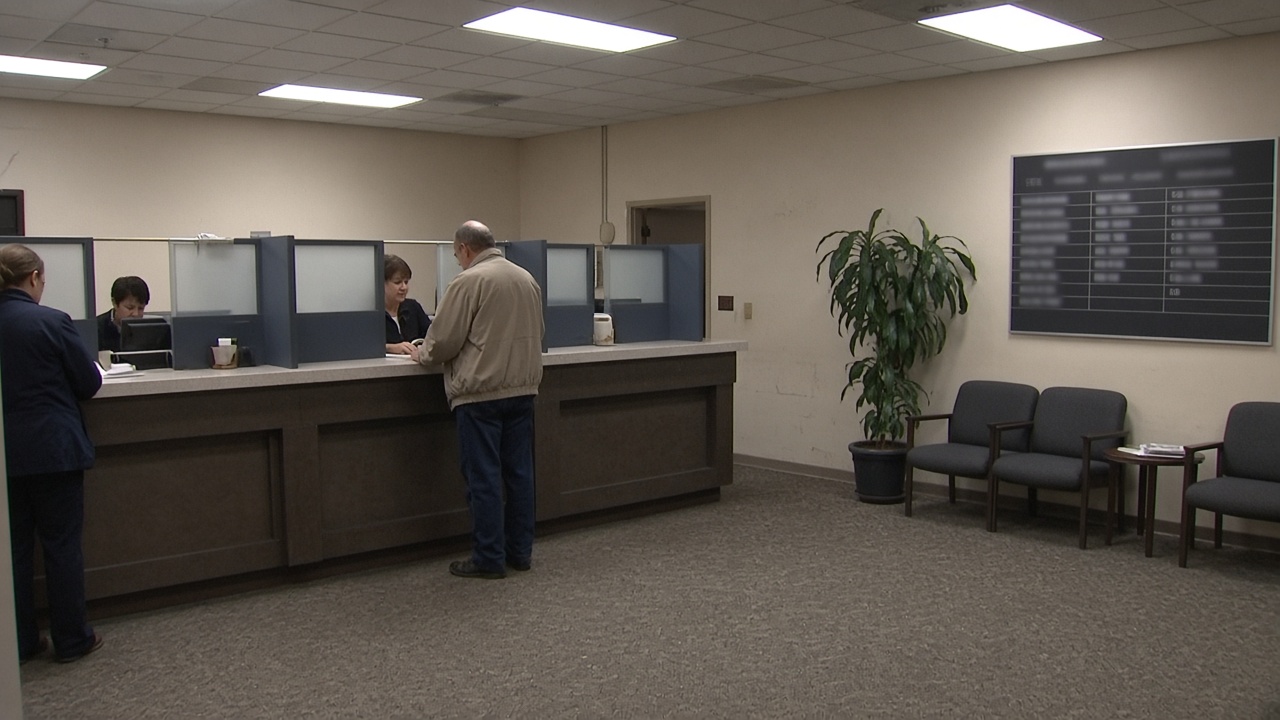 Lobby of a modest credit union branch with members at a teller counter and a wall-mounted rate board, fluorescent ceiling panels, worn carpet, no logos or readable text