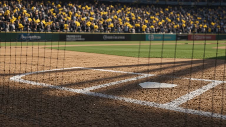 Baseball stadium infield dirt and chalk lines seen from behind home plate netting, yellow fan towels blurred in stands, afternoon shadows, no logos or readable text