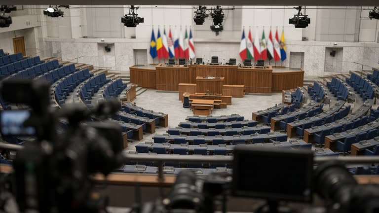 National Assembly plenary chamber seen from the press gallery, empty blue seats and central podium with microphones, overhead cameras, flags blurred, no readable signage.