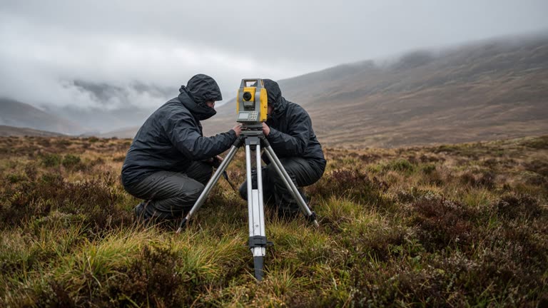 Scientists in outdoor weather jackets adjusting a tripod-mounted survey instrument on Scottish moorland grass, low cloud on hills, wind-blown heather, documentary field photo, no faces shown clearly